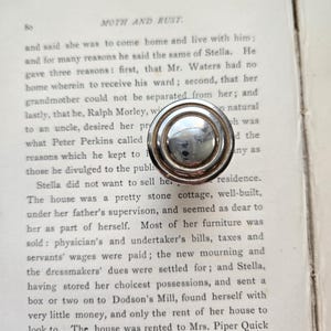 May include: A silver-toned, circular drawer pull with a raised, concentric design, resting on an open book. The book's pages display printed text, with the title "MOTH AND RUST" visible at the top. The pull has a reflective surface.