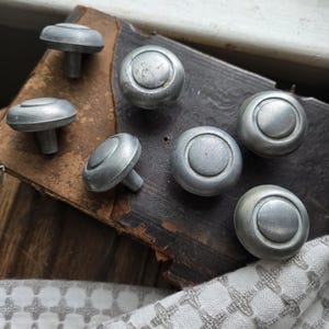 May include: A collection of six antique silver-toned cabinet knobs. The knobs have a mushroom-shaped design with a circular face and a stem. They are arranged on a dark, textured surface with a patterned white and gray fabric in the foreground.