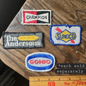 May include: A collection of vintage embroidered patches featuring various brand names and logos. Patches include "Champion," "The Andersons," "Sunoco," and "Sohio." The patches are rectangular and oval, with a wooden ruler for scale.