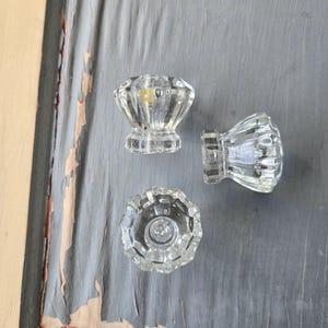 May include: Three clear glass cabinet knobs with a faceted design. The knobs are arranged on a weathered gray surface. The text "sold individually" is at the bottom of the image.