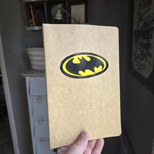 Pode incluir: Um caderno retangular com uma capa de cor castanho-claro ou bege. Centrado na capa frontal encontra-se um log&oacute;tipo do Batman oval e proeminente, com uma silhueta de morcego preta sobre um fundo amarelo brilhante com um rebordo preto.