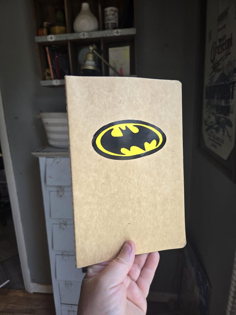 Pode incluir: Um caderno retangular com uma capa de cor castanho-claro ou bege. Centrado na capa frontal encontra-se um log&oacute;tipo do Batman oval e proeminente, com uma silhueta de morcego preta sobre um fundo amarelo brilhante com um rebordo preto.