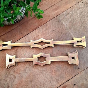 May include: Two antique brass drawer pulls with a decorative diamond-shaped center and flared ends. The pulls are a warm gold color and rest on a wooden surface. A small green plant in a silver pot is in the background.