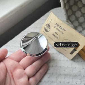 May include: A shiny, chrome-colored die-cast knob, 2 inches in diameter, with a reflective surface. The knob is held in a hand, with a vintage label visible in the background. The word "vintage" is written on the image.