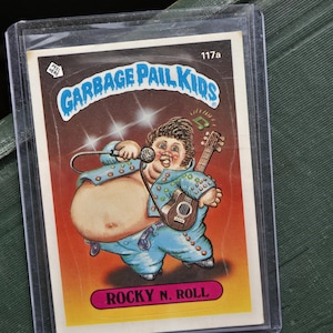 May include: A Garbage Pail Kids trading card featuring a cartoon character in a blue jumpsuit, holding a microphone and a small guitar. The card is titled "Rocky N. Roll" and is encased in a protective sleeve. The card has a brown and orange background.