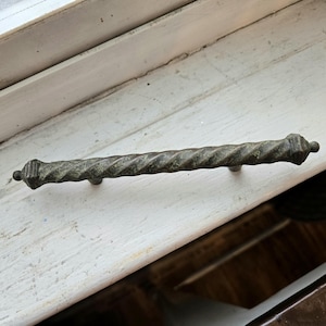 May include: A weathered, antique-style drawer pull with a twisted, rope-like design. The metal hardware has a dark, aged finish and decorative end caps. The pull is mounted on a white surface.