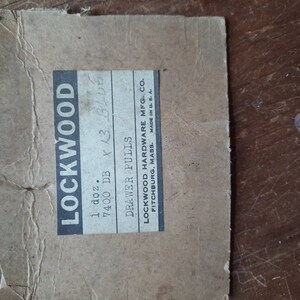 Puede incluir: Una caja de cart&oacute;n marr&oacute;n con una etiqueta blanca que dice "LOCKWOOD 1 doz. 7400 DB x 13 BLUE DRAWER PULLS LOCKWOOD HARDWARE MFG. CO. FITCHBURG, MASS. MADE IN U.S."