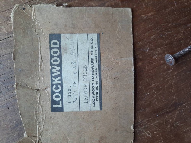 Puede incluir: Una caja de cart&oacute;n marr&oacute;n con una etiqueta blanca que dice "LOCKWOOD 1 doz. 7400 DB x 13 BLUE DRAWER PULLS LOCKWOOD HARDWARE MFG. CO. FITCHBURG, MASS. MADE IN U.S."