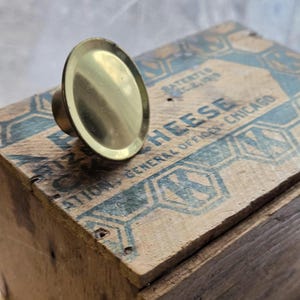 May include: A brass-colored cabinet knob with a circular, flat face and a short, cylindrical stem. The knob is set against a weathered wooden surface with faded blue text that reads "HEESE" and "GENERAL OFFICES - CHICAGO".