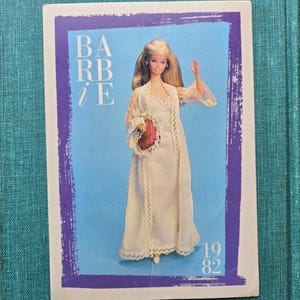 May include: A vintage Barbie doll image from 1982. The doll is wearing a white dress with lace trim and a matching robe. The image has a blue background with the word "Barbie" in purple letters.