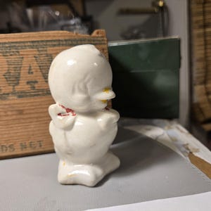 May include: A white ceramic figurine of a bird with a red bow and yellow beak. The bird is standing on a flat surface.