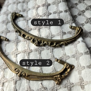 May include: Two antique brass drawer pulls, labeled "style 1" and "style 2". Style 1 features ornate scrollwork, while style 2 has a simpler design. Both are curved with decorative end pieces. The background is a patterned fabric.