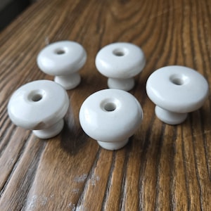 May include: A set of five white ceramic knobs, each featuring a smooth, rounded top surface with a centered circular hole. These hardware pieces have a glossy finish and a tapered base, designed for attachment to furniture or cabinetry doors.