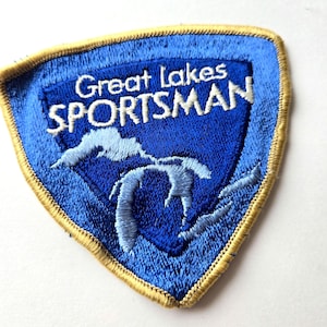 May include: Embroidered patch with a blue shield design featuring the text "Great Lakes SPORTSMAN" in white. The patch has a gold border and depicts a light blue outline of the Great Lakes. The patch is a vintage style.