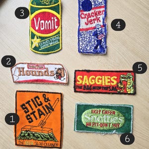 May include: Six embroidered iron-on patches with various designs and slogans. The patches are on a light brown wooden surface. The patches include "Vomit", "Cracker Jerk", "Hounds", "SAGGIES THE BAG WITH THE SAG", "STIC & STAIN THE BIG SLOB CLEANER", and "UGLY GREEN SNOTTIES NO FLY-DON'T TRY".