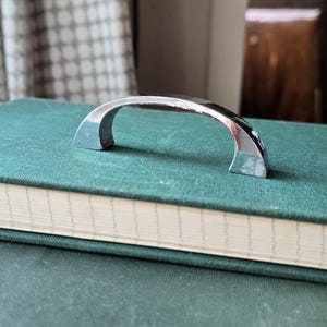 May include: A shiny, chrome-finished cabinet pull handle with a curved design. The handle is resting on a stack of two green books. The background includes a patterned curtain and a wooden cabinet.