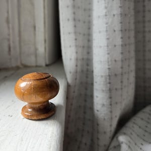 May include: A small, wooden mushroom-shaped cabinet knob with a light brown finish. The knob has a rounded top with a carved design and a flared base. It sits on a white surface near a window.