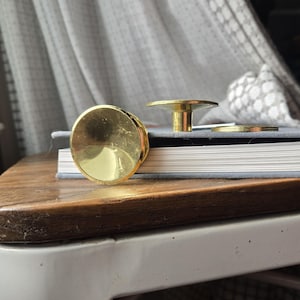 May include: Close-up of several gold-colored cabinet knobs and pulls. One large, round knob is in the foreground, with a smaller, mushroom-shaped pull and a thin ring pull behind it. The hardware sits on a stack of books on a wooden table.