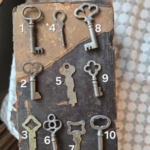 May include: A collection of ten antique keys of various designs, including heart, clover, and oval shapes. The keys are made of metal and are arranged on a worn, dark brown surface. The keys are numbered 1 through 10.