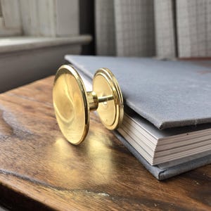 Può includere: Un pomello circolare dorato con uno stelo, appoggiato su una superficie di legno accanto a un libro grigio. Il pomello ha una superficie lucida e riflettente. Il libro ha una copertina grigia e pagine bianche.