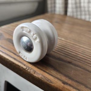 May include: A white porcelain cabinet knob with a silver metal center. The knob has a rounded shape with a circular design and is set against a wooden surface.