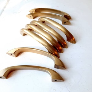 May include: A set of eight gold-toned cabinet door pulls. The pulls are all the same size and shape, and they have a slightly curved design. They are arranged in a stack, with the top pull slightly offset from the others.