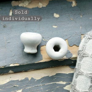 May include: Two white ceramic knobs with a smooth, rounded profile. One knob is shown from the side, while the other is viewed from the top, revealing a hexagonal center opening. The text "Sold individually" is visible in the upper left corner.