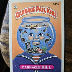 May include: A Garbage Pail Kids trading card featuring "Barnacle Bill." The card shows a cartoon character with a fishbowl for a head, filled with fish and a miniature castle. The character wears a blue sailor suit. The card has the text "GARBAGE PAIL KIDS" at the top.