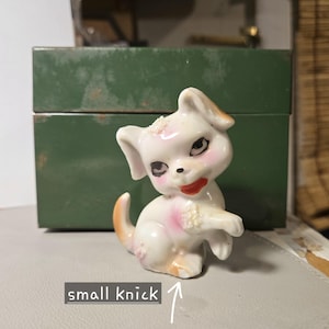 May include: White porcelain figurine of a dog with pink and brown accents. The dog has a playful expression and is sitting with its paw raised. There is a small nick on the figurine's side.