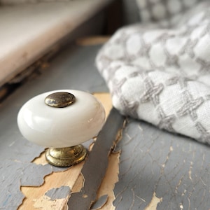 May include: A round, white ceramic cabinet knob featuring a smooth, glossy finish. The center of the knob is accented with a small, circular, distressed brass-toned metal cap, and the base is constructed from matching distressed brass-toned metal.