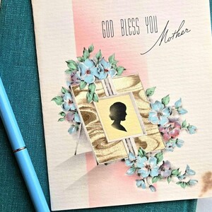 May include: Vintage Mother's Day card with a silhouette in a wooden frame, surrounded by blue and pink flowers. The card reads "GOD BLESS YOU Mother" and is accompanied by a light blue pen.