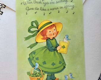 Pode incluir: Cartão de felicitações vintage do Dia de São Patrício com uma menina num vestido verde e chapéu amarelo, segurando um livro e rodeada por pássaros azuis. O cartão tem o texto "When Irish eyes are smiling..." num fundo verde claro.