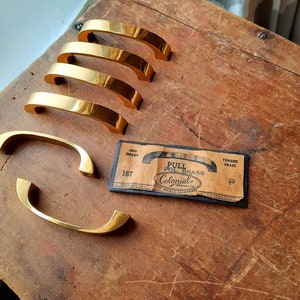 May include: Five gold-toned cabinet pulls with a curved design. The pulls are arranged in a stack on a wooden surface. A small card with the text "Pull Pol Brass Colonial Bronze Co. Torrington, Conn. Forged Brass" is visible in the image.