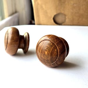 May include: Two wooden cabinet knobs with a warm brown finish. One knob is a rounded shape with a circular design, and the other is a more traditional knob shape. The knobs are set against a white background.