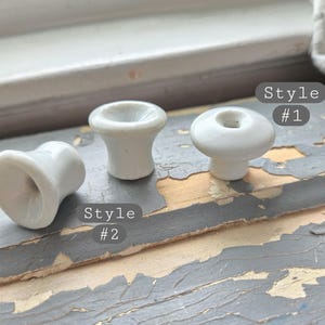 May include: Three white ceramic knobs with smooth, glossy finishes. Two knobs are shaped like flared cylinders, labeled Style #2, and one knob is shaped like a rounded disc with a central indentation, labeled Style #1, all featuring a central hole.