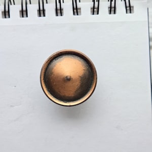 May include: A copper-colored, round knob with a conical center. The knob has a dark, aged patina around the edges, giving it a vintage appearance. The knob is likely for a drawer or cabinet.