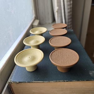 May include: Eight cabinet knobs, four are brown and four are beige. The knobs have a textured surface and a raised center. The knobs are arranged in two rows of four on a dark green book.