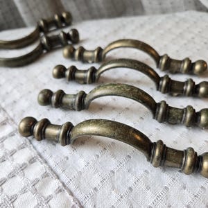 May include: Several antique-style bronze drawer pulls with a curved handle design. The pulls have a distressed finish, giving them a vintage appearance. They are arranged on a white, textured surface.
