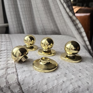 May include: A collection of five brass cabinet knobs. Four knobs have a rounded, ball-like design, while the fifth has a mushroom-shaped top. The knobs are displayed on a white, textured surface.