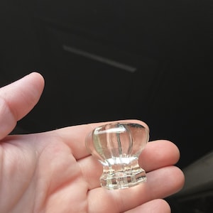May include: A clear glass cabinet knob with a rounded top and a detailed base. The knob is held in a hand against a dark background. The knob has vertical ridges and a decorative design.