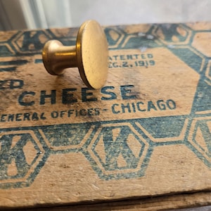 May include: A vintage brass stamp with a circular head and a short handle rests on a wooden box. The box has blue and white geometric designs and the words "CHEESE" and "CHICAGO" printed on it. The stamp is likely used for marking or sealing.