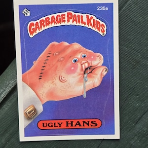 May include: A Garbage Pail Kids trading card featuring a grotesque hand with stitches, blue eyes, and drool. The card has the text "GARBAGE PAIL KIDS" and "UGLY HANS" on a blue background.