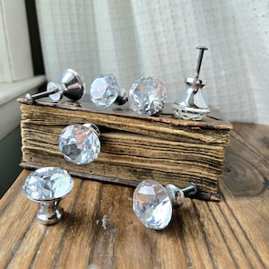 Puede incluir: Colección de pomos y tiradores de cristal transparente y facetado con herrajes plateados. Los pomos se exhiben sobre un libro antiguo y una superficie de madera, mostrando su diseño decorativo y brillante. Ideales para añadir un toque de elegancia a los muebles.