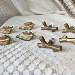 May include: A collection of decorative cabinet knobs in leaf and branch designs. The knobs are made of a metallic gold material and are mounted on white hardware. The knobs are arranged on a white patterned surface.