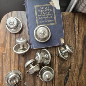 May include: A collection of vintage metal cabinet knobs with a distressed finish, scattered on a wooden surface. A small, worn blue book titled "God's Minute" is also present. The knobs have a classic, industrial design.