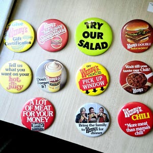 May include: Nine colorful Wendy's restaurant promotional buttons with various slogans and images, including a hamburger, salad, chili, and a pick-up window.