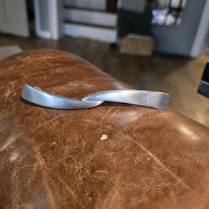 May include: Two brushed silver cabinet handles with a curved design. The handles are connected and appear to be new. The handles are resting on a brown leather surface.