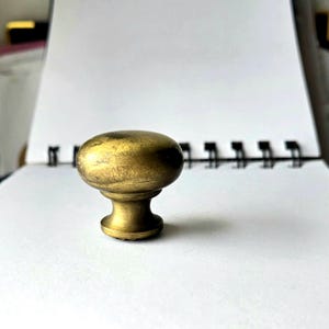 May include: A vintage-style brass cabinet knob with an oval top and a short, rounded base. The knob has a warm, golden hue and a slightly aged appearance, suggesting it is an antique or a reproduction. It is designed for use on drawers or cabinet doors.