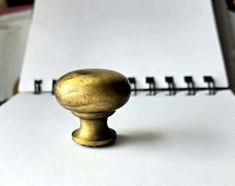 Pode incluir: Um puxador de armário de latão em estilo vintage com um topo oval e uma base curta e arredondada. O puxador tem um tom dourado quente e uma aparência ligeiramente envelhecida, sugerindo que é antigo ou uma reprodução.