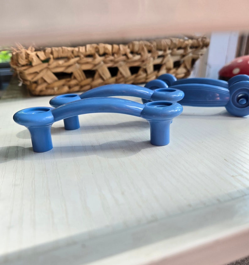 Puede incluir: Dos pomos de armario de pl&aacute;stico azul con un dise&ntilde;o curvo. Los pomos tienen un acabado liso y brillante.